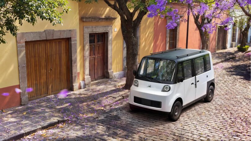 Prototipo del Olinia Uno blanco con techo negro estacionado sobre una calle empedrada de un pueblo mexicano con fachadas coloridas y flores de jacaranda